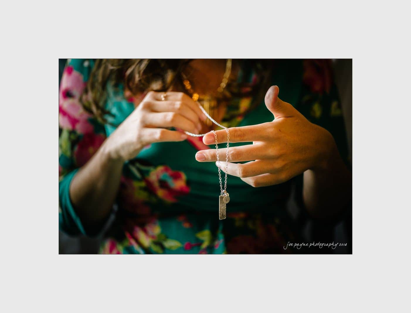 wilmington wedding photographer - nichole & eric A person wearing a floral dress holds a necklace with a pendant in both hands. The focus is on the hands and necklace, with soft lighting highlighting the jewelry.