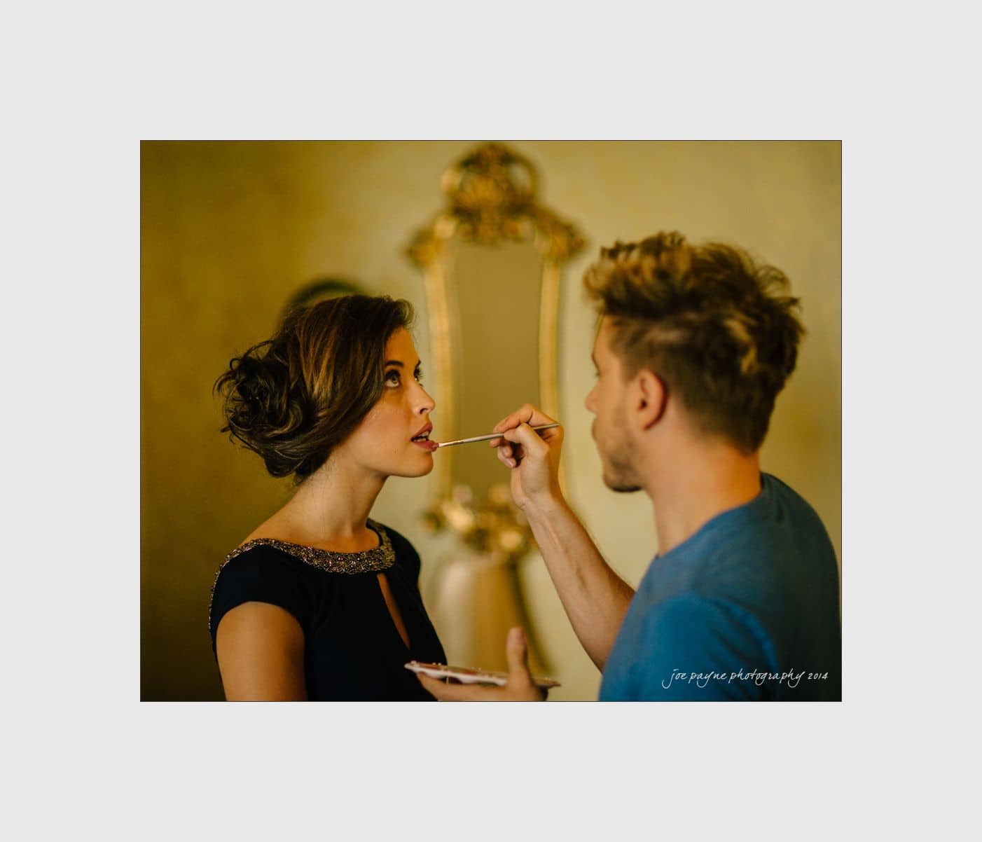 wilmington wedding photographer - nichole & eric A makeup artist applies lipstick to a woman with dark hair, who is wearing a black dress. The setting appears elegant, with a gold-framed mirror in the background.
