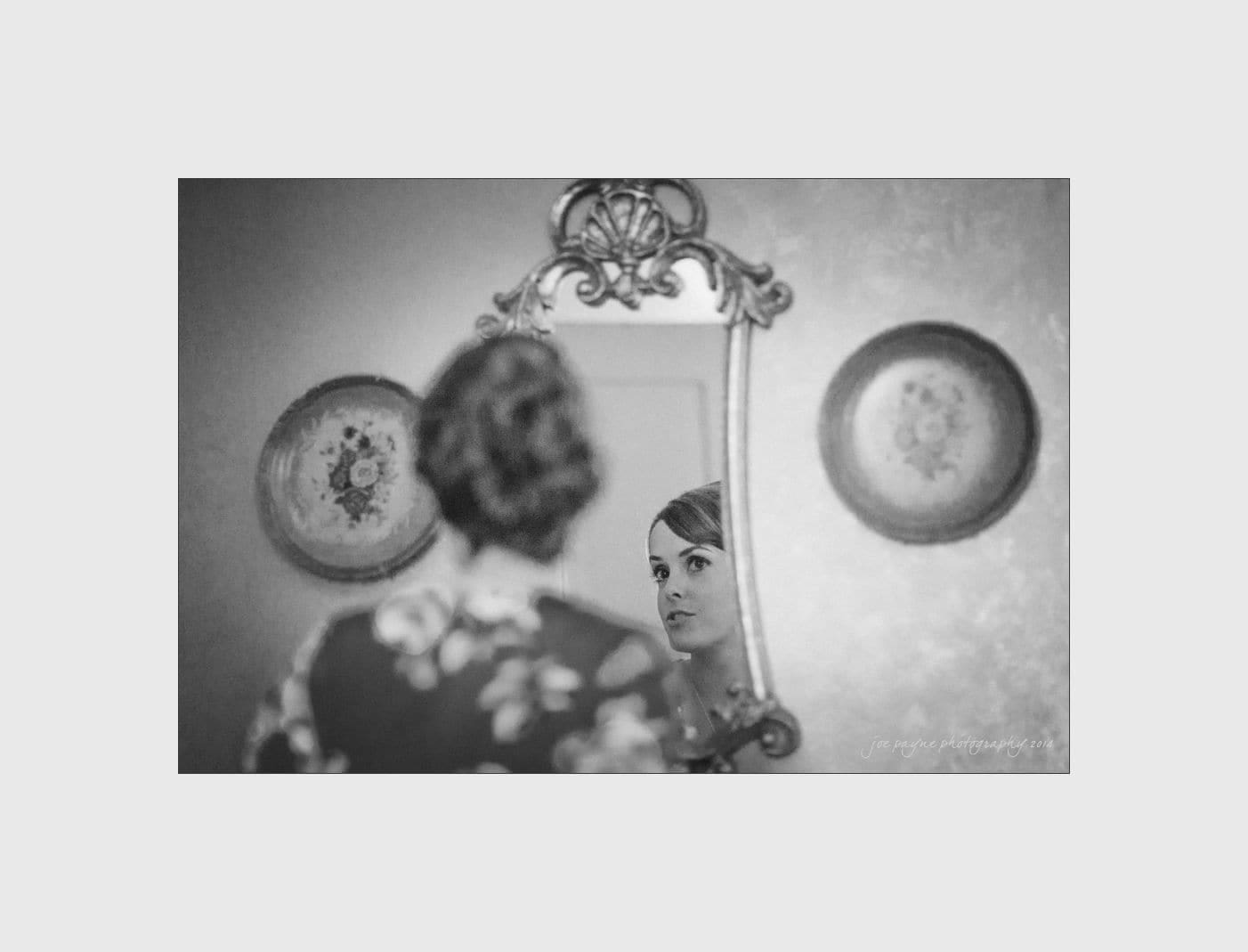 wilmington wedding photographer - nichole & eric A woman is looking at her reflection in an ornate mirror on the wall. The room is softly lit, with decorative plates hanging nearby. The woman has a calm, contemplative expression. The photo is in black and white.