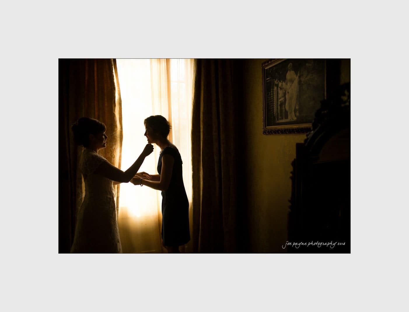 wilmington wedding photographer - nichole & eric A silhouette of two women stands by a window with sheer curtains, one adjusting the others collar. The room is dimly lit, with a framed painting on the wall. A warm, intimate ambiance is captured.