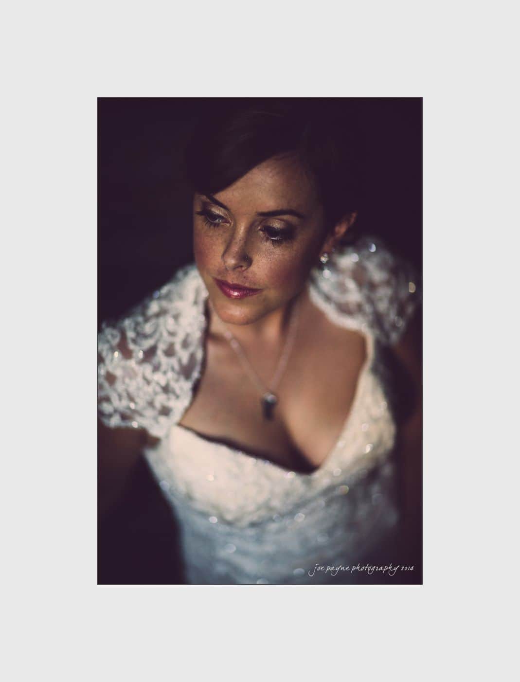 wilmington wedding photographer - nichole & eric A woman in a white lace wedding dress gazes downward, captured in soft, dim lighting. She wears a necklace and her hair is styled up. The photo has a dark, moody atmosphere.