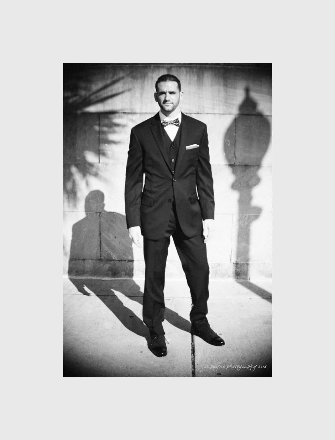 wilmington wedding photographer - nichole & eric A man in a formal suit and bow tie stands on a sunlit sidewalk. His shadow is visible on the ground. The background includes a stone wall and a vintage street lamp casting a shadow. The image is in black and white.