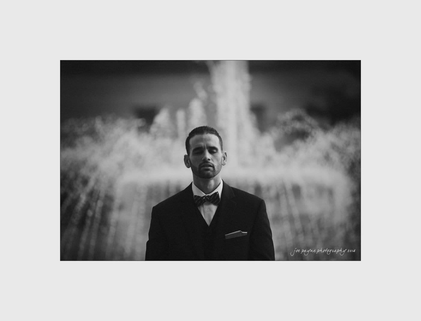 wilmington wedding photographer - nichole & eric Black and white photo of a man in a suit standing in front of a large, spray-style fountain. He has a serious expression and the background is blurred, emphasizing the water and his formal attire.