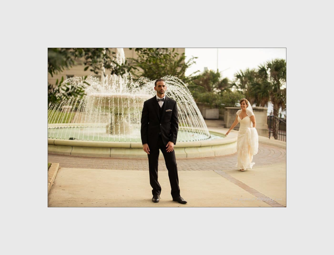 wilmington wedding photographer - nichole & eric A groom in a black suit stands in front of a fountain, looking forward, while a bride in a white gown walks towards him from behind. Palm trees and a building are visible in the background.
