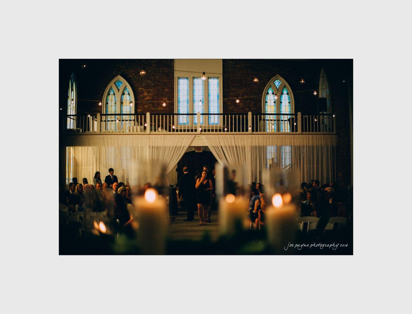 wilmington wedding photographer - nichole & eric A dimly lit wedding venue with candles in the foreground. Guests are seated on either side, and a woman stands in the aisle. Stained glass windows and draped curtains add elegance to the setting.