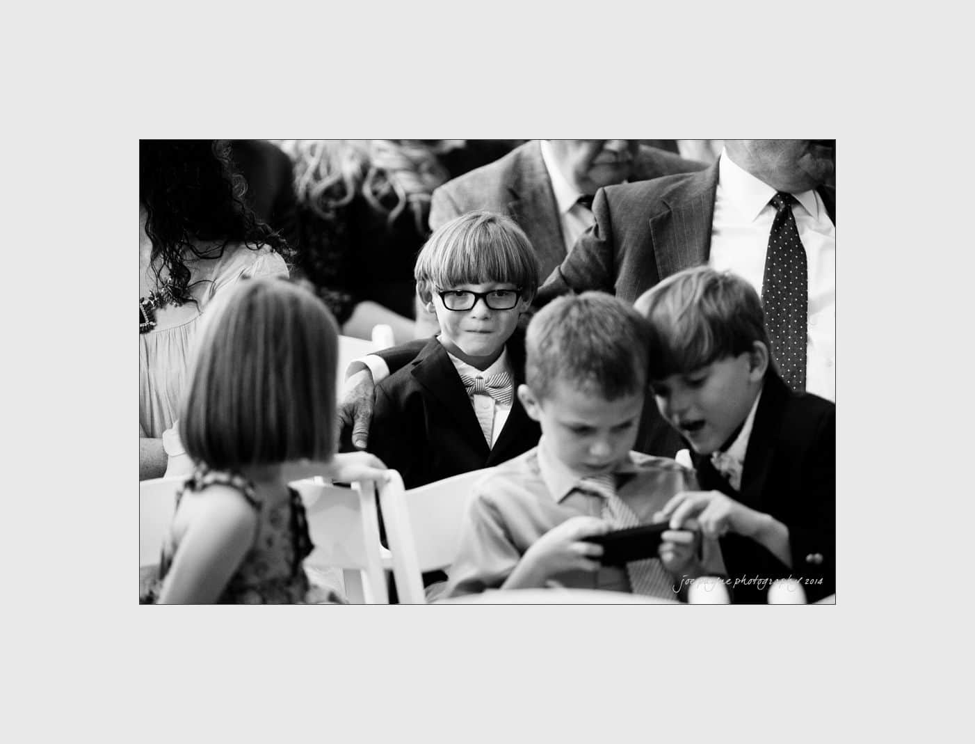 wilmington wedding photographer - nichole & eric A black and white photo of a group of young children sitting together. One boy with glasses looks at the camera, while others are focused on a device. The setting appears formal, with people in suits and dresses around them.