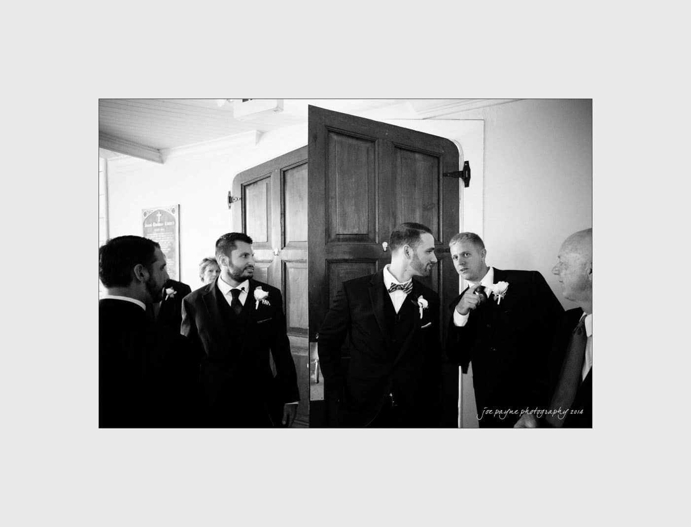 wilmington wedding photographer - nichole & eric A group of men in suits are gathered near a wooden door. One man leans in to whisper to another, who is standing in the doorway. They all appear to be in conversation in a bright room, and each wears a boutonniere.