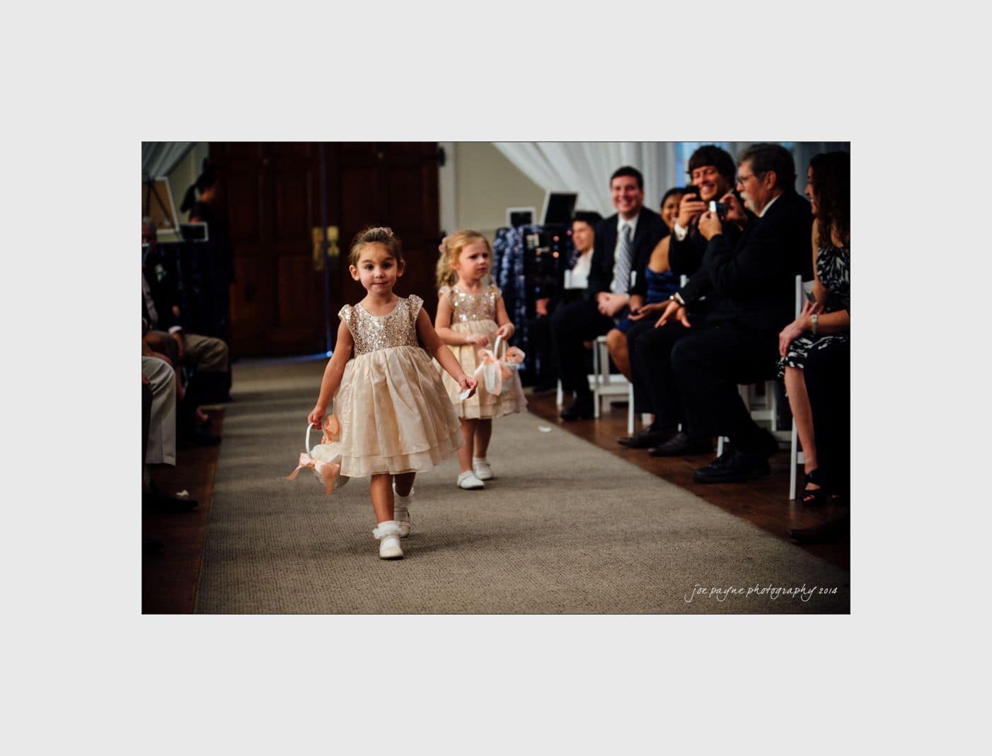 wilmington wedding photographer - nichole & eric Two young flower girls in matching lace dresses walk down an aisle, holding flower petals. They are followed by a crowd of smiling guests seated on either side, capturing the moment with their phones and cameras.
