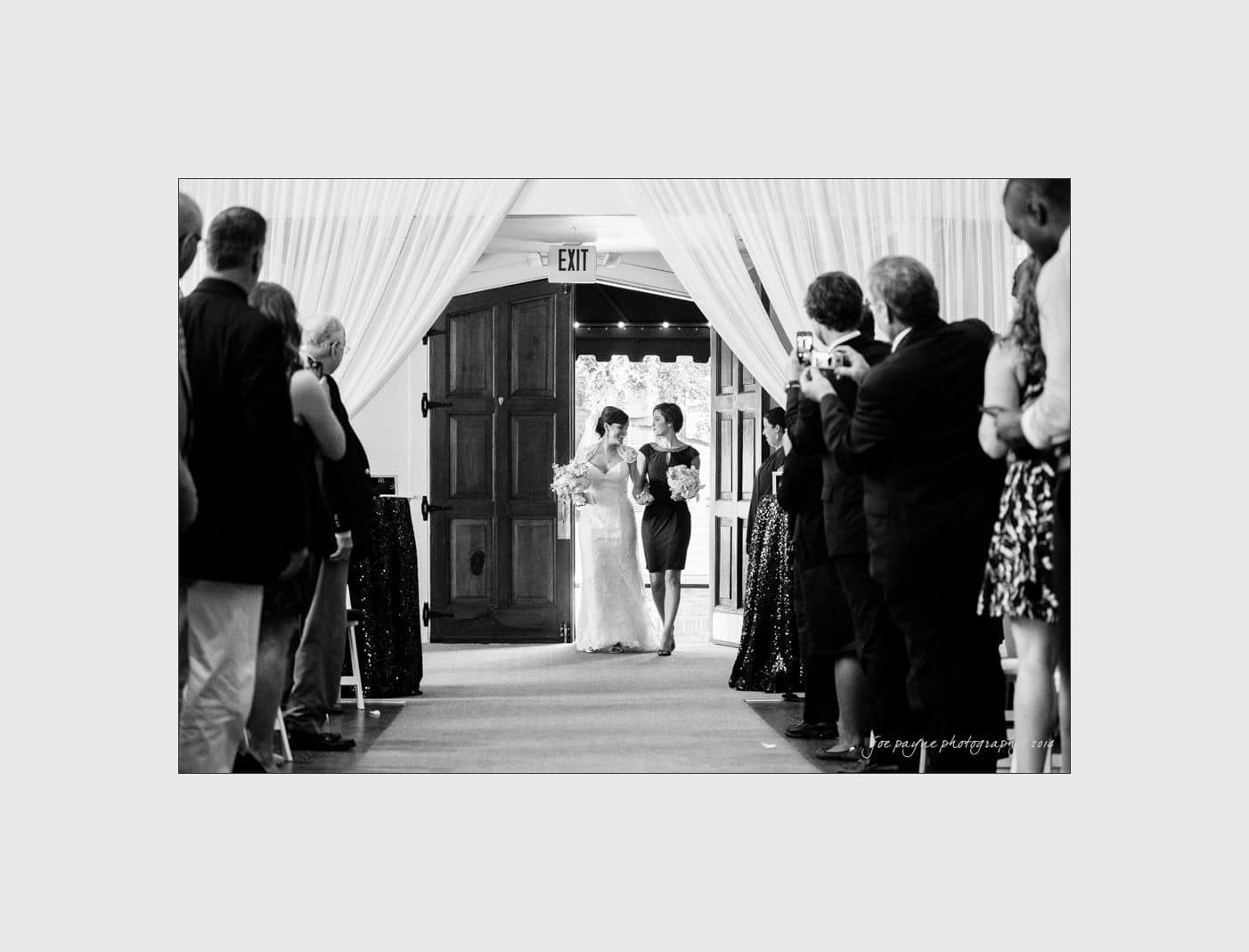 wilmington wedding photographer - nichole & eric A bride walks down the aisle with two companions, surrounded by guests in formal attire who are taking photos. The scene is indoors with a doorway in the background, framed by draped curtains. The image is in black and white.