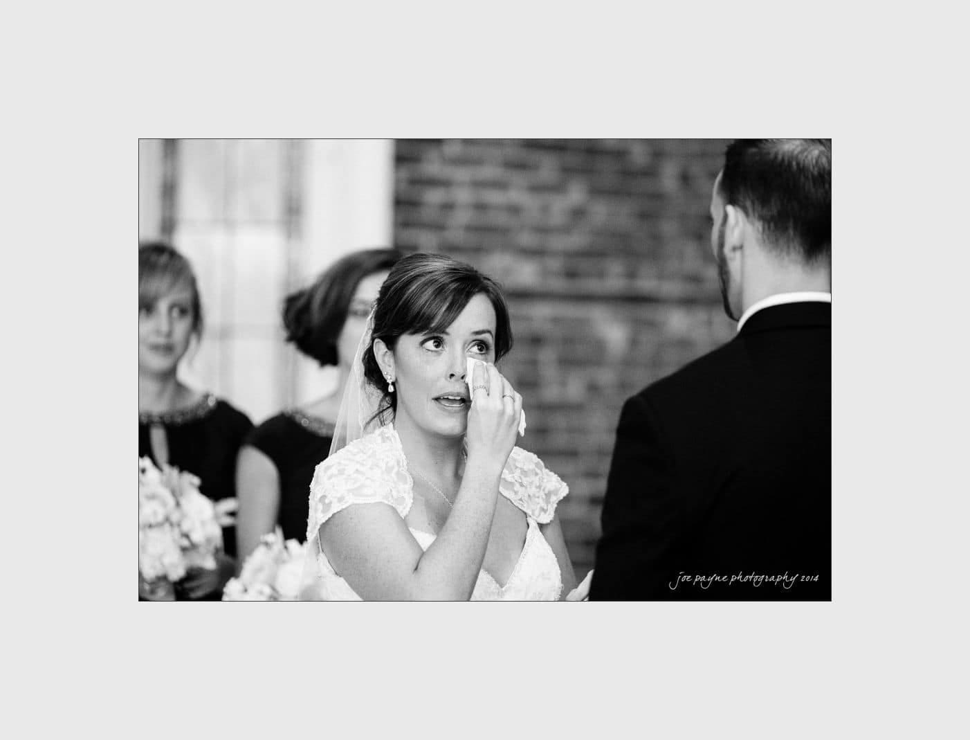 wilmington wedding photographer - nichole & eric A bride in a wedding dress wipes her eye during a ceremony, standing across from a groom in a suit. Bridesmaids holding bouquets are visible in the background. The setting is indoors with a brick wall.