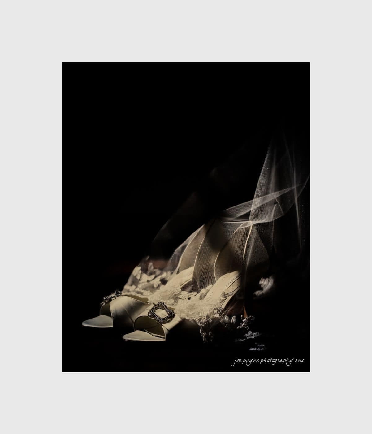 wilmington wedding photographer - nichole & eric A pair of elegant white high-heeled shoes with satin and lace details sit on a dark background. A sheer veil with lace edges partially covers the shoes, creating a soft, dreamy effect.