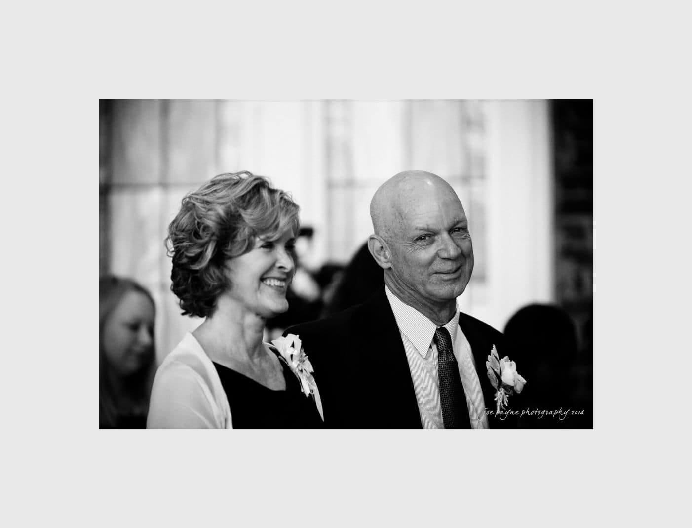wilmington wedding photographer - nichole & eric A black and white photo of a smiling woman and an older man wearing suits and corsages, standing together. They appear to be at a formal event, such as a wedding, with people seated in the background.
