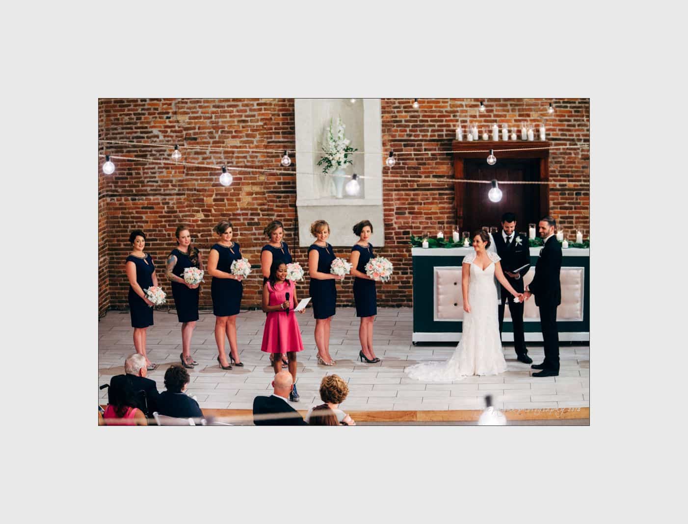 wilmington wedding photographer - nichole & eric A wedding ceremony with a bride and groom holding hands. Bridesmaids in matching navy dresses hold bouquets, and a young girl in a pink dress stands among them. The venue has a brick wall and decorative lights hanging above.