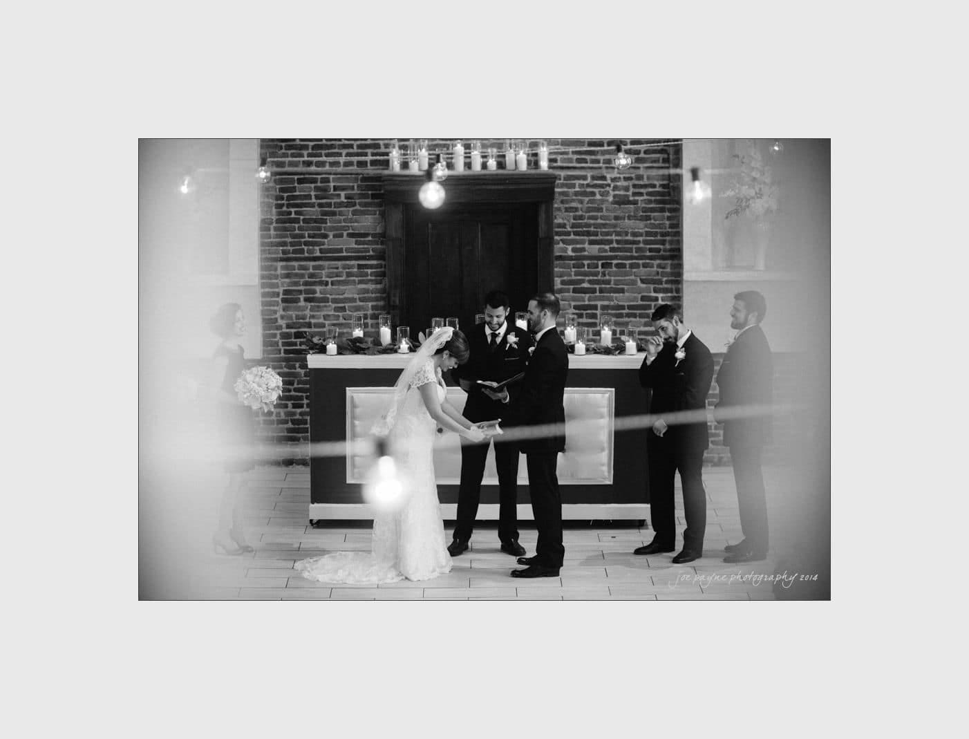 wilmington wedding photographer - nichole & eric A black and white photo of a wedding ceremony. The bride and groom, holding hands, stand in front of an officiant. They are in an elegant setting with brick walls and soft lighting, flanked by two attendants.
