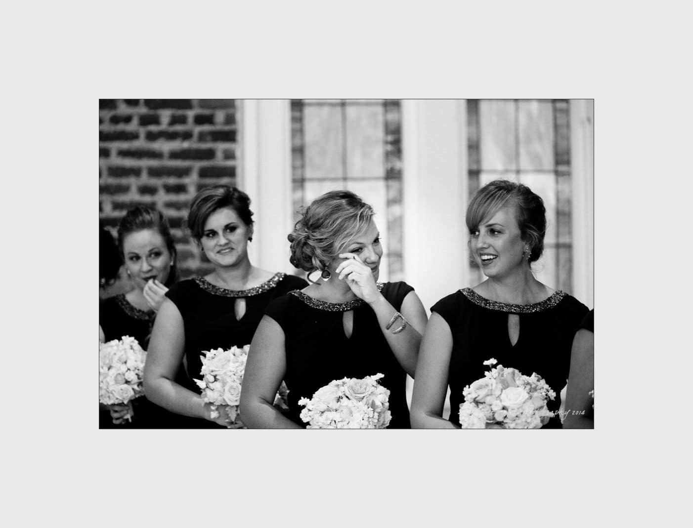 wilmington wedding photographer - nichole & eric Four bridesmaids in matching dresses, holding bouquets, smile and laugh during a wedding ceremony. The background features a brick wall and large windows, creating a warm and joyful atmosphere.