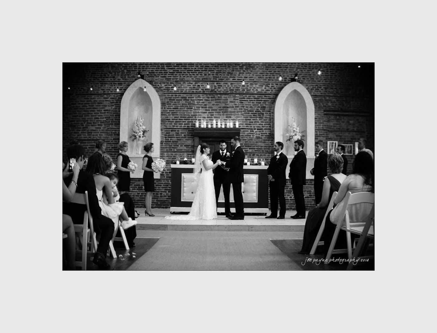 wilmington wedding photographer - nichole & eric A black and white photo of a wedding ceremony in a church. The bride and groom stand at the altar holding hands. Bridesmaids and groomsmen are beside them, and guests are seated. Candles and floral arrangements decorate the background.