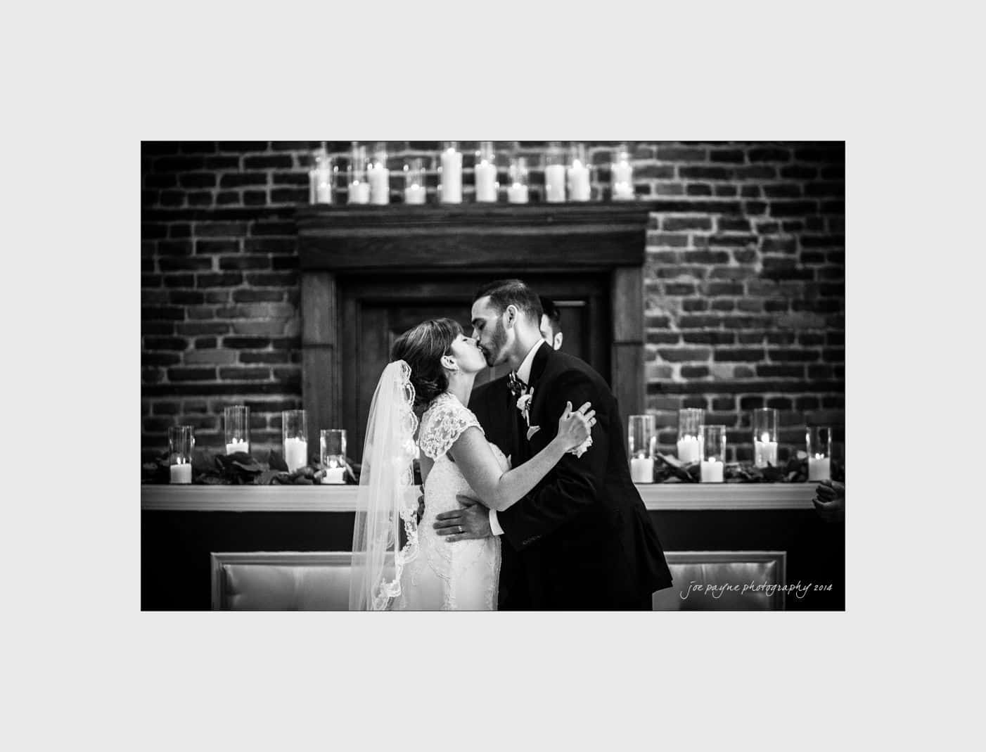 wilmington wedding photographer - nichole & eric A couple shares a kiss during their wedding ceremony. They stand in front of a rustic brick wall adorned with lit candles. The bride wears a lace gown and veil, and the groom is in a formal suit. The image is in black and white.