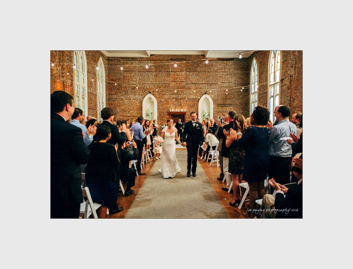 wilmington wedding photographer - nichole & eric A newlywed couple walks down the aisle hand in hand in a brick-walled venue, smiling as guests stand and applaud. The couple is dressed in a wedding gown and suit. String lights and arched windows adorn the background.