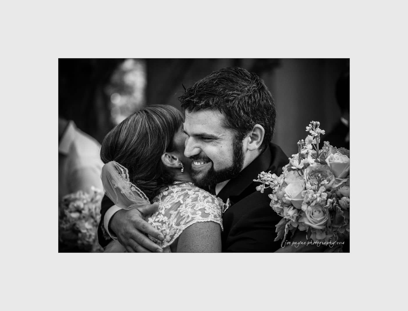 wilmington wedding photographer - nichole & eric A black and white image of a joyful groom hugging a bride who is holding a bouquet. The groom has a wide smile, and the brides lace dress and floral arrangement are visible. Both appear to be celebrating, possibly at a wedding ceremony.