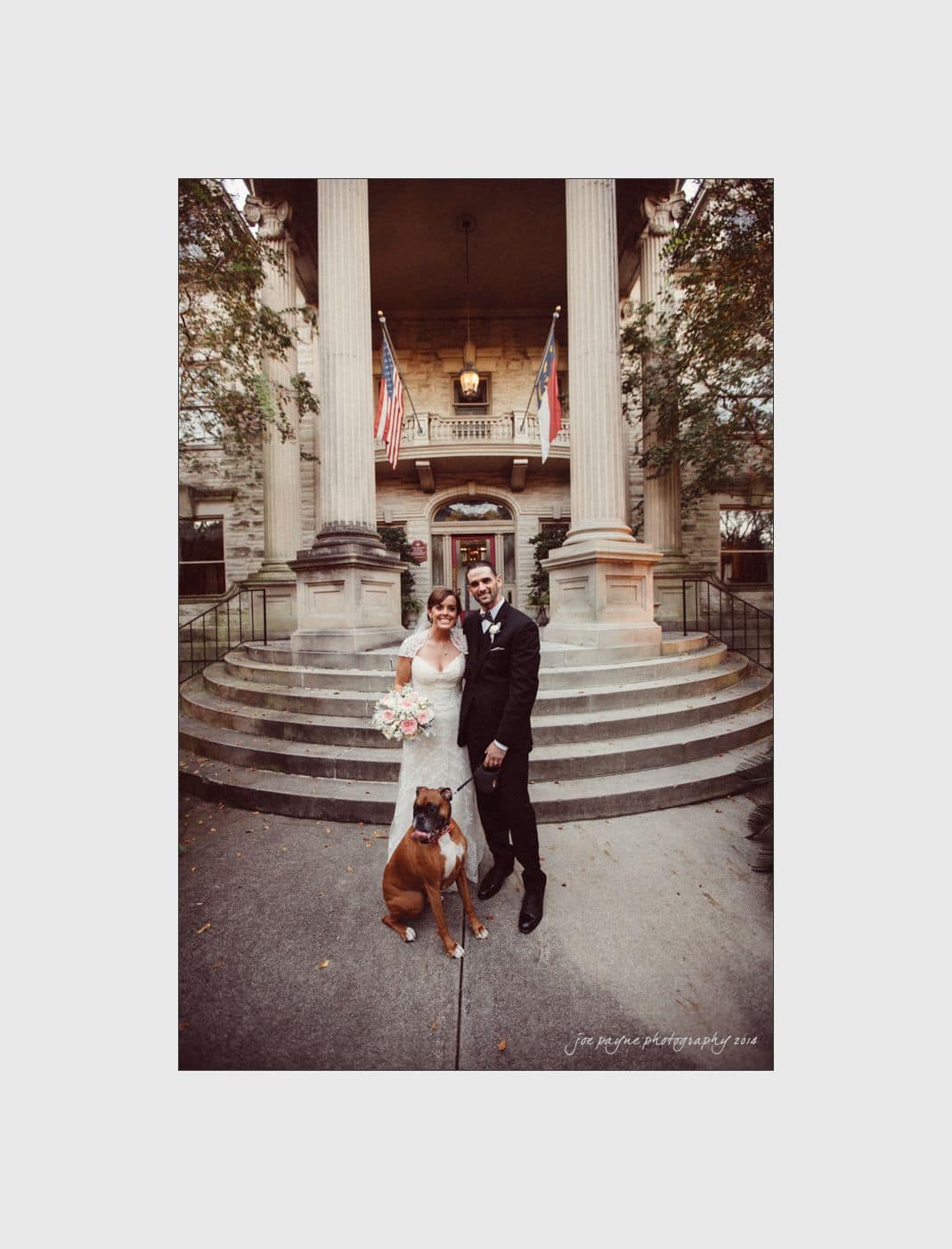wilmington wedding photographer - nichole & eric A bride and groom stand in front of a grand building with columns, accompanied by a brown dog. The bride holds a bouquet of flowers. Two flags are visible in the background. The scene has a romantic and formal atmosphere.