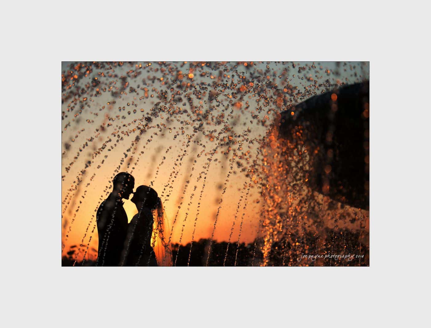 Silhouetted couple embracing behind a fountain with water droplets glistening. The sky in the background is painted with warm hues of orange and pink as the sun sets.