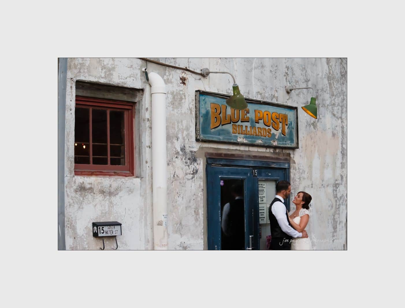 wilmington wedding photographer - nichole & eric A couple stands outside a vintage building with a weathered exterior. The sign reads Blue Post Billiards. They are dressed in wedding attire, with the bride in a white gown and the groom in a vest and tie, sharing an intimate moment.