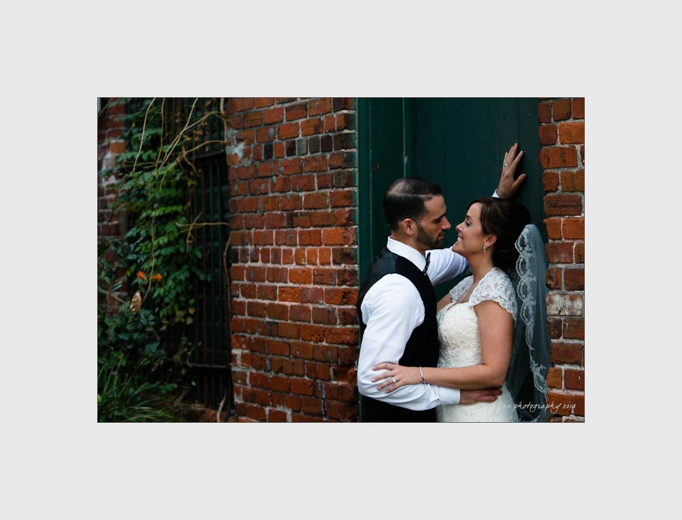 wilmington wedding photographer - nichole & eric A bride and groom embrace and gaze into each others eyes while standing against a brick wall. The bride wears a lace gown and veil, and the groom is in a vest and tie. Green foliage peeks out beside the wall, adding a touch of nature.