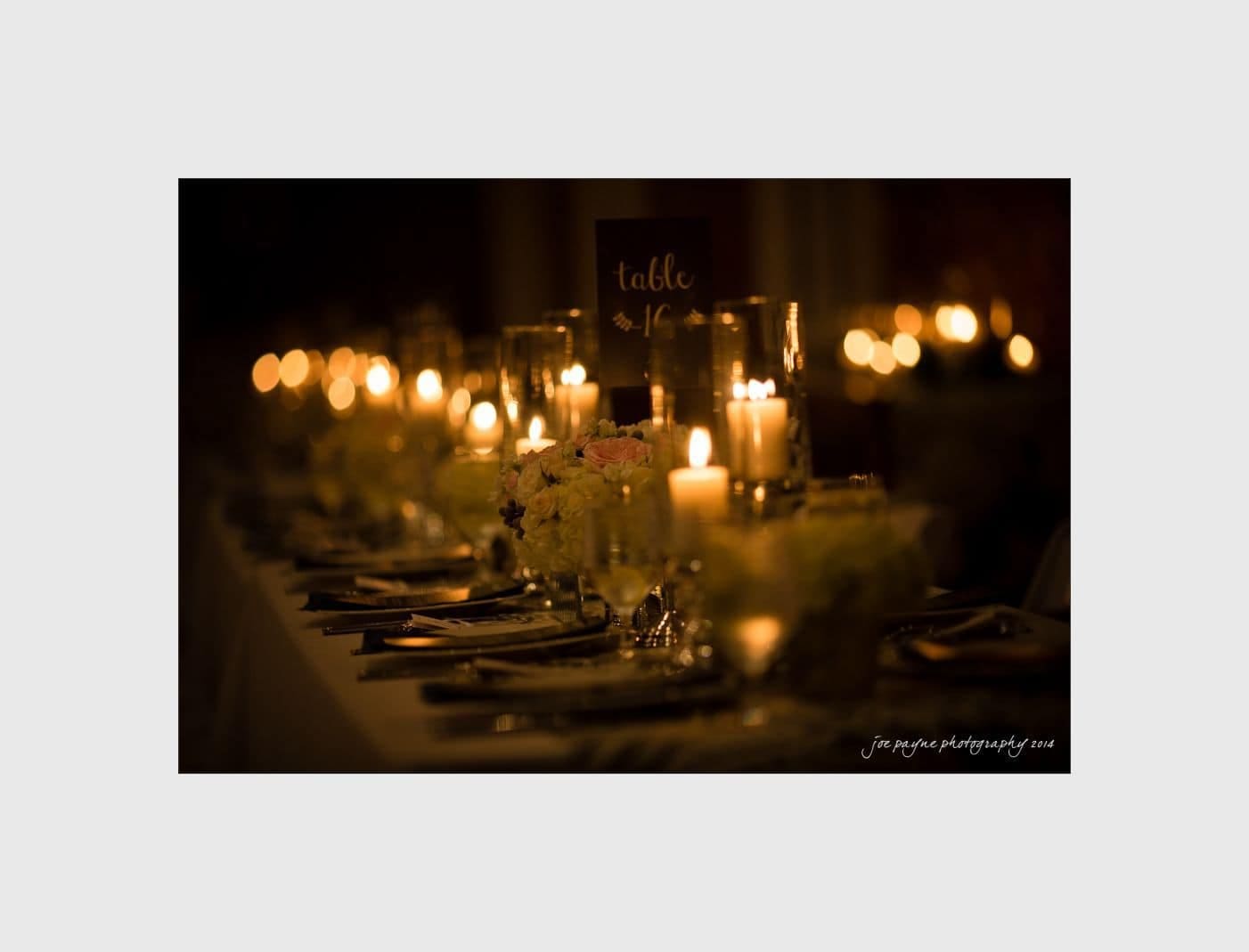 wilmington wedding photographer - nichole & eric A dimly lit banquet table decorated with white flowers, candles, and elegantly set dinnerware. The warm glow from the candles creates a cozy and inviting atmosphere. A sign reading table 1 is visible among the decorations.