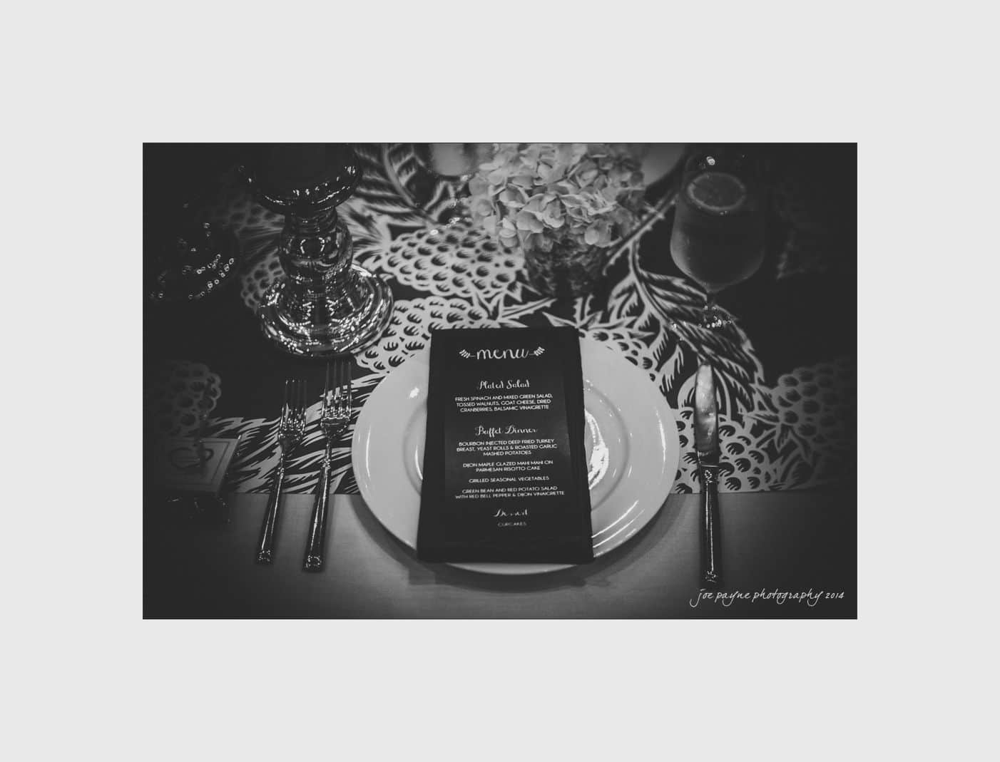 wilmington wedding photographer - nichole & eric A black and white photo of an elegantly set dining table. A white plate is centered with a dark menu placed on top. Cutlery is arranged neatly on either side. Decorative patterns and a glass are visible in the background.