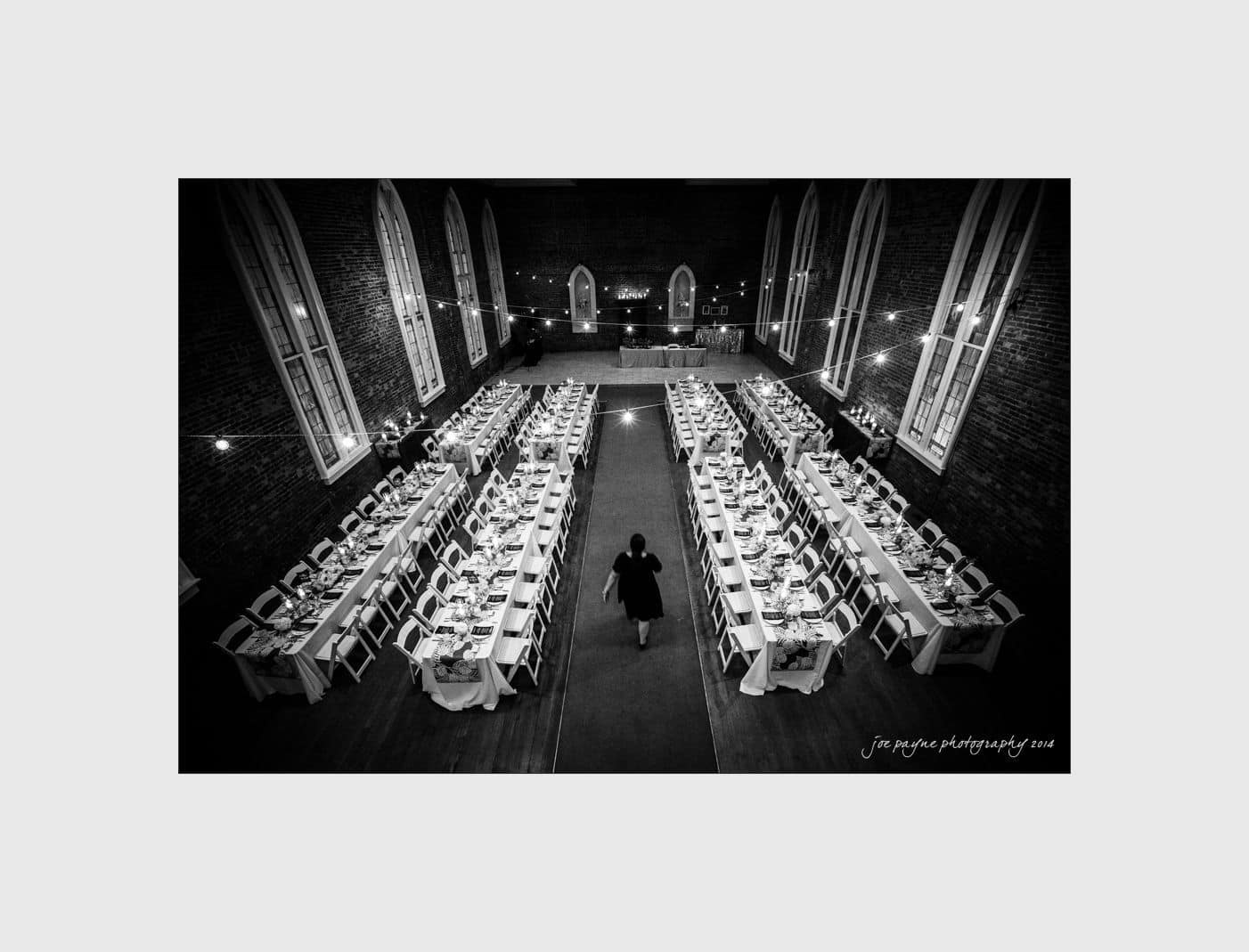 wilmington wedding photographer - nichole & eric Black and white photo of a large dining hall with long tables set for a formal event. A person walks down the center aisle between the tables. Tall, arched windows line the brick walls.