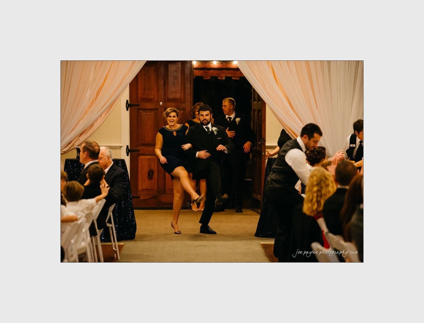wilmington wedding photographer - nichole & eric A couple energetically enters a room at a wedding reception, dancing with high kicks. Guests seated at tables watch and smile. The room is decorated with draped curtains and wooden doors. Two men in suits stand in the background.
