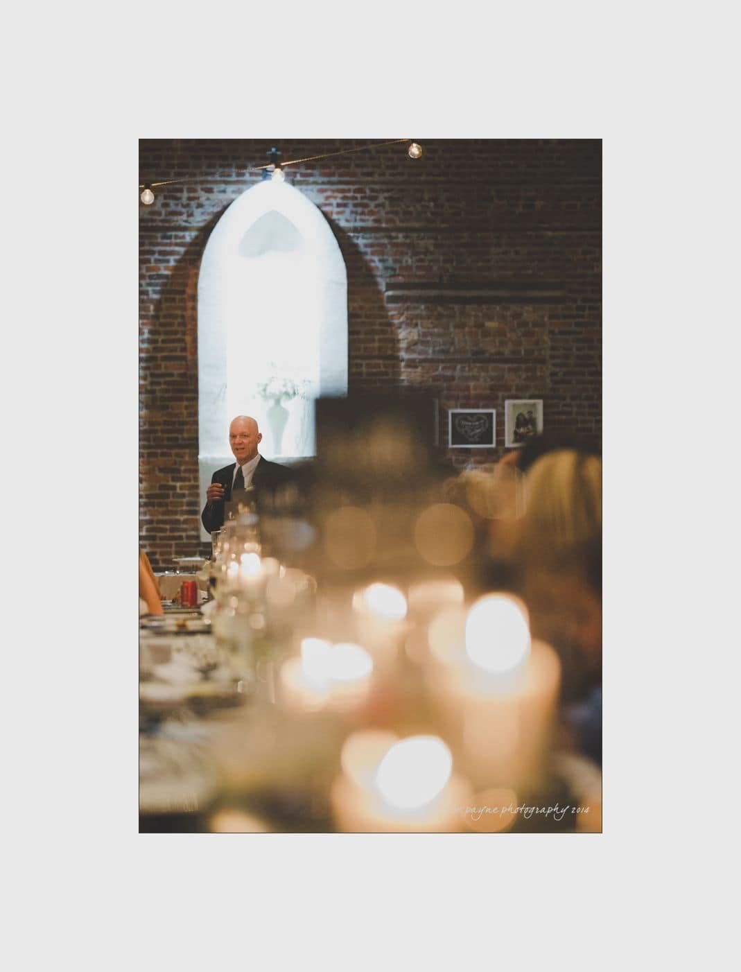 wilmington wedding photographer - nichole & eric A person stands in front of a brick wall with a large arched window, addressing an audience in a dimly lit room. Soft candle lights create a warm atmosphere, with blurry figures seated in the foreground.