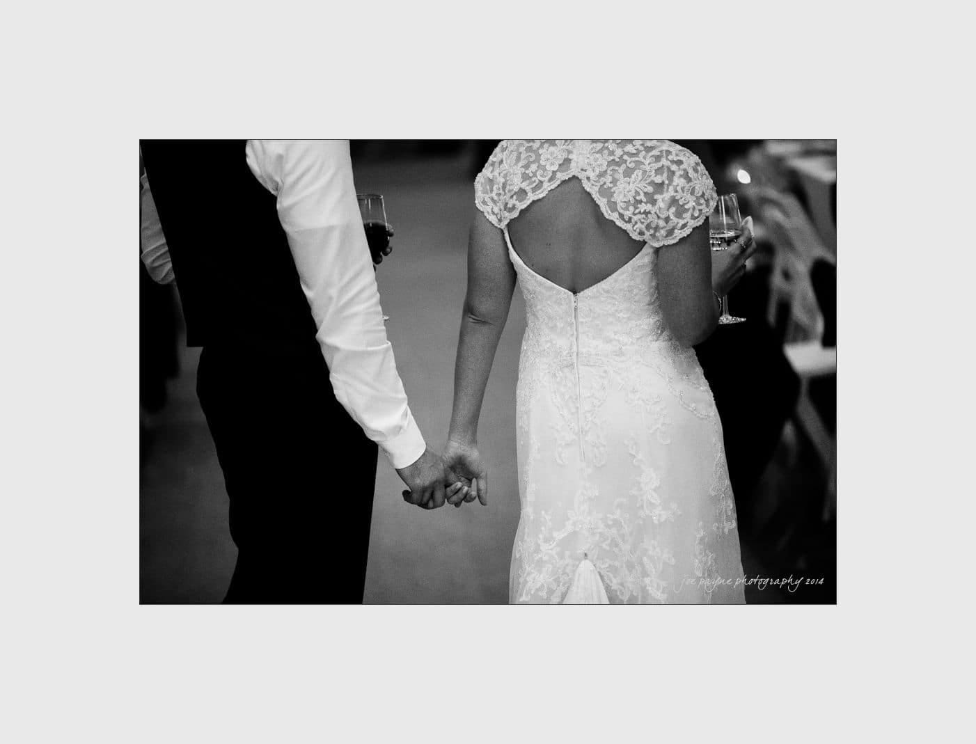 wilmington wedding photographer - nichole & eric A black and white photo of a couple holding hands at a wedding. The woman is wearing a lace wedding dress with an open back, and the man is in a suit vest. They are facing away, captured mid-step.