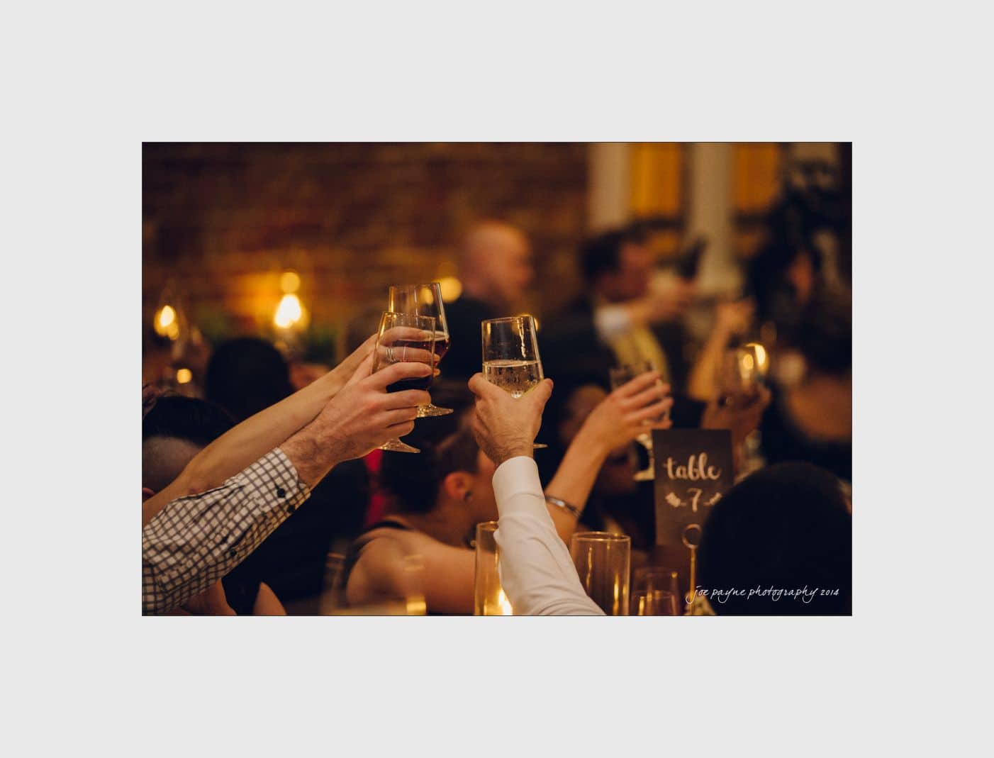 wilmington wedding photographer - nichole & eric People raise glasses in a toast at a warmly lit gathering. A table 7 sign is visible, indicating a celebratory event.