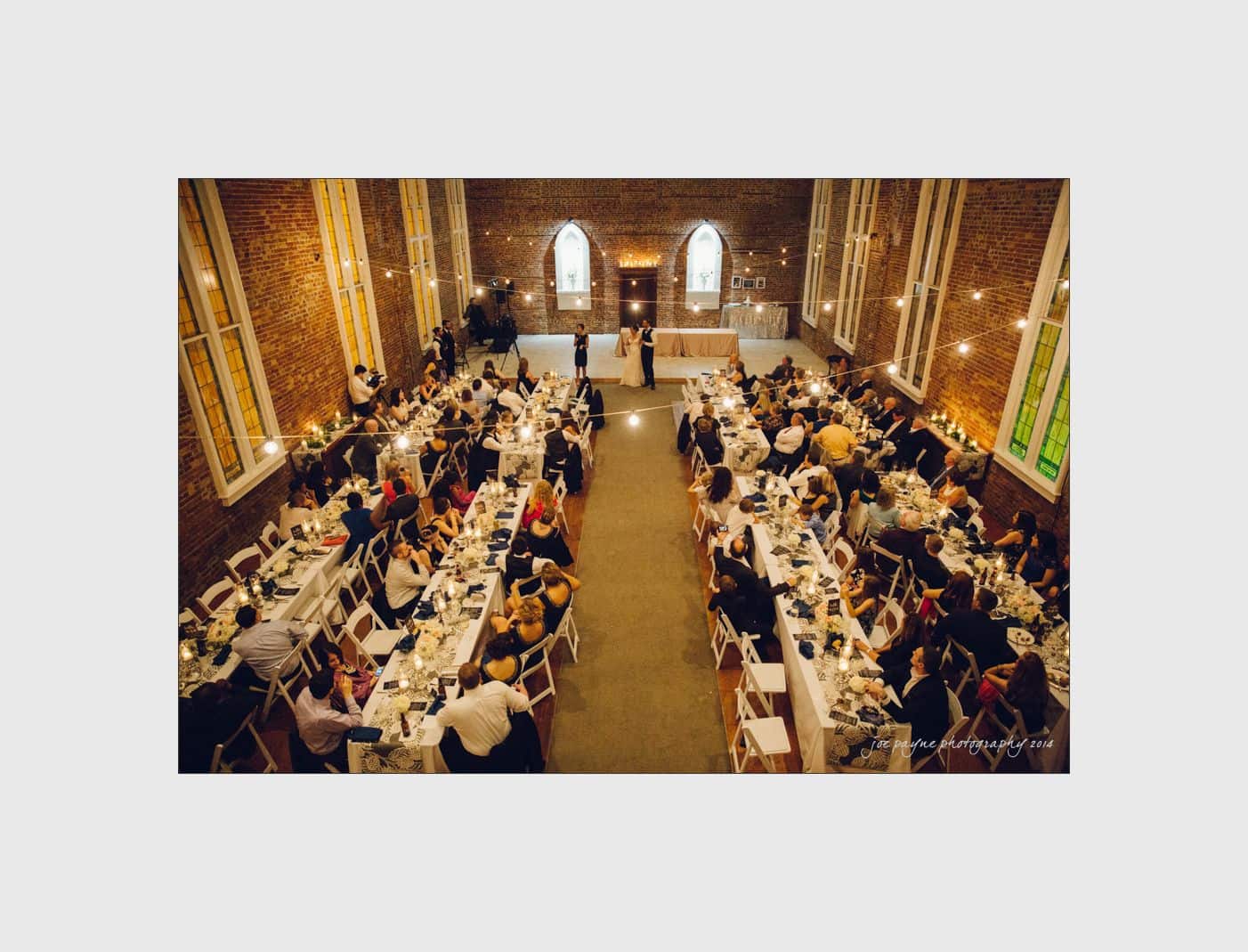 wilmington wedding photographer - nichole & eric A large wedding reception in a grand hall with high ceilings and brick walls. Guests are seated at long tables adorned with white tablecloths and floral centerpieces. The bride and groom are dancing in the center under warm lights.