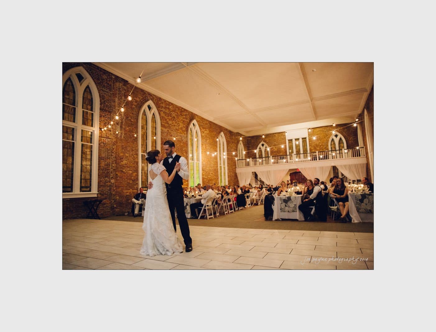 wilmington wedding photographer - nichole & eric A couple dances in a spacious, elegantly decorated hall with large windows and brick walls. Guests sit at tables adorned with white tablecloths, watching the couple. Warm lighting creates a romantic ambiance.