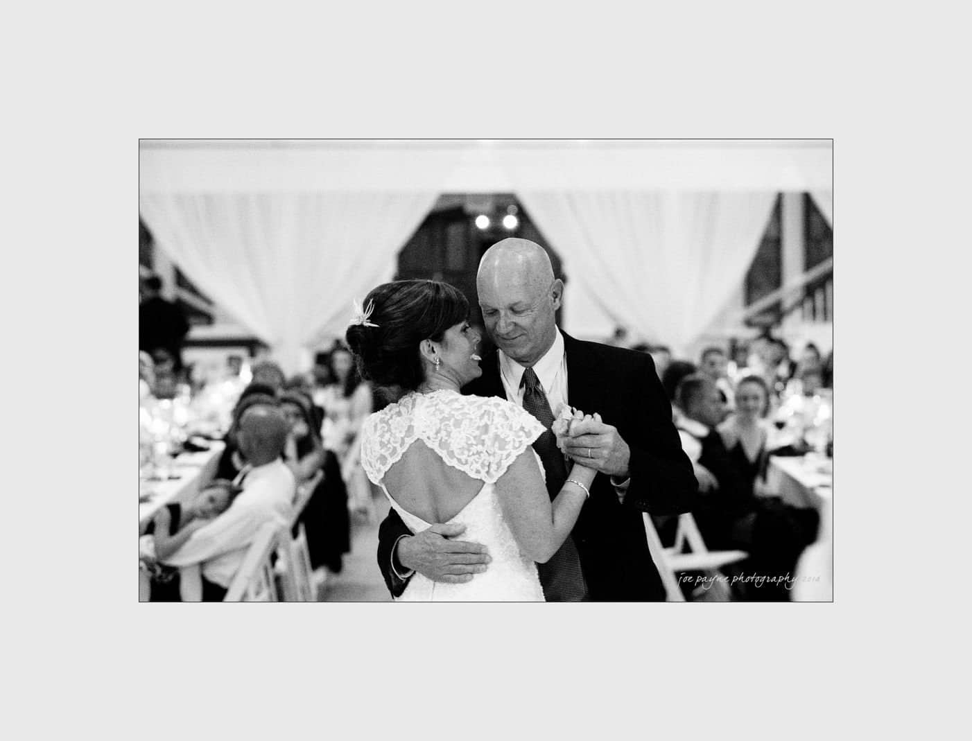wilmington wedding photographer - nichole & eric A bride and an older man, possibly her father, share a dance in a warmly lit reception hall. They are surrounded by seated guests and white drapery, creating an elegant atmosphere.