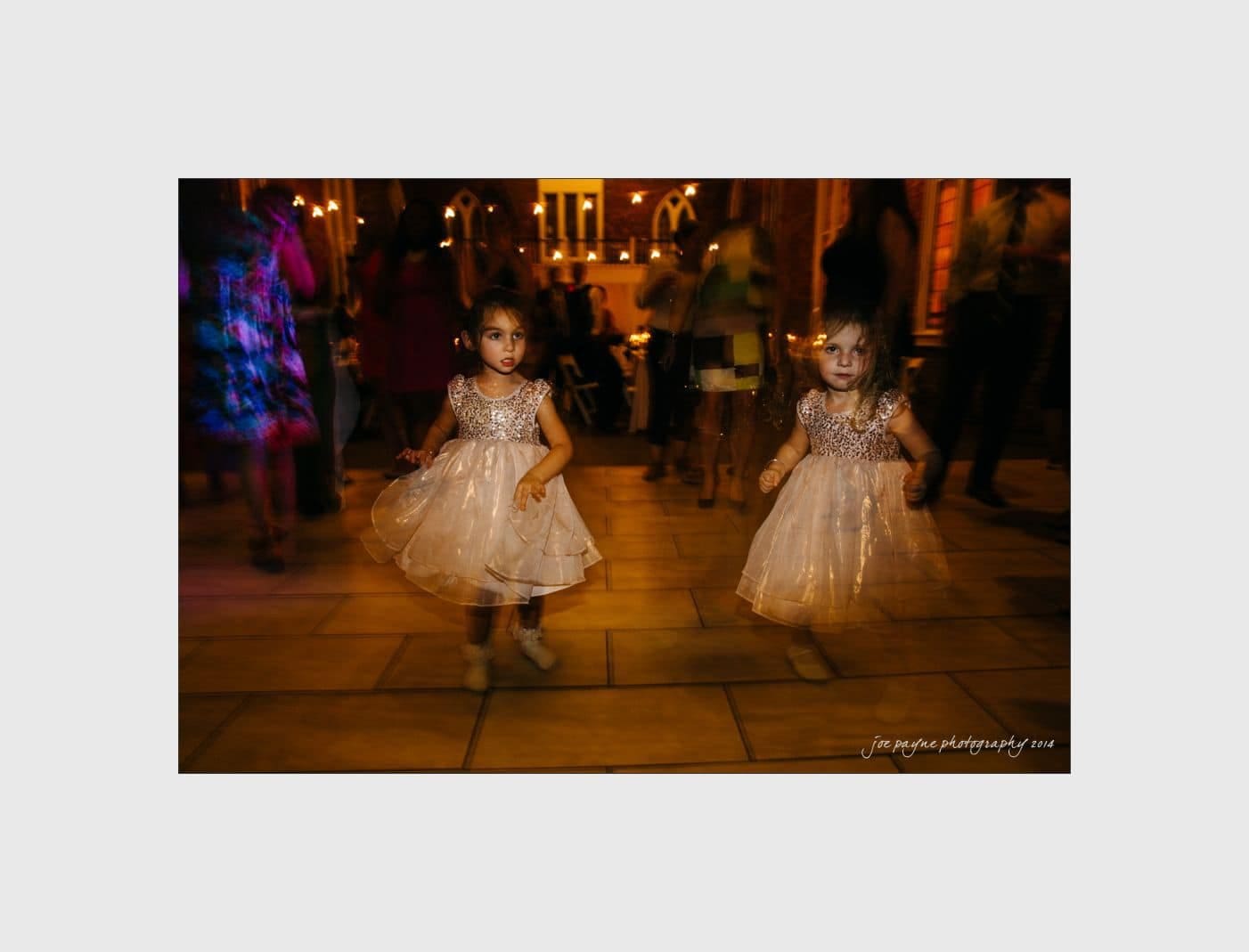 wilmington wedding photographer - nichole & eric Two young girls in sparkly dresses are standing on a dimly lit dance floor. The background shows blurred silhouettes of people dancing, with warm lighting creating a festive atmosphere.