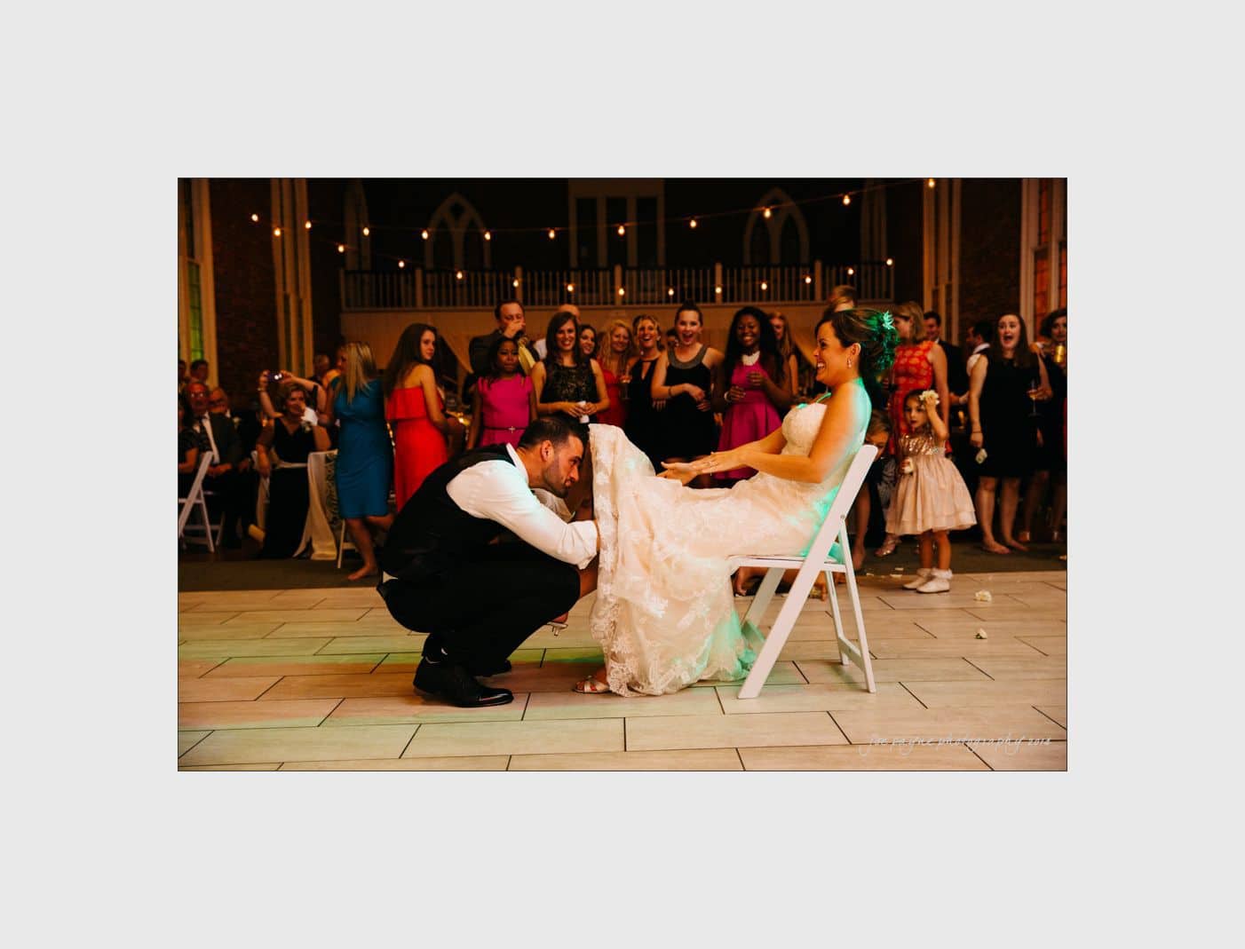 wilmington wedding photographer - nichole & eric A groom playfully removes a garter from the brides leg in a lively wedding reception scene. The bride is seated in a chair, smiling and wearing a white dress. Guests in colorful outfits watch and laugh in the background.