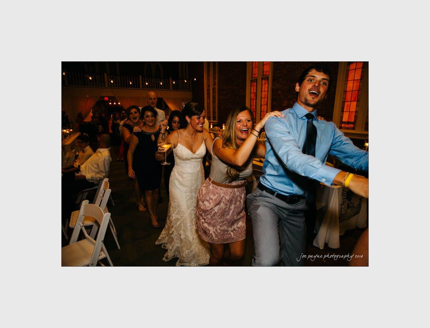wilmington wedding photographer - nichole & eric A group of people, led by a man in a blue shirt and tie, are joyfully dancing in a conga line at a wedding reception. The bride, wearing a white dress, smiles as she holds a glass. The atmosphere is festive and lively.