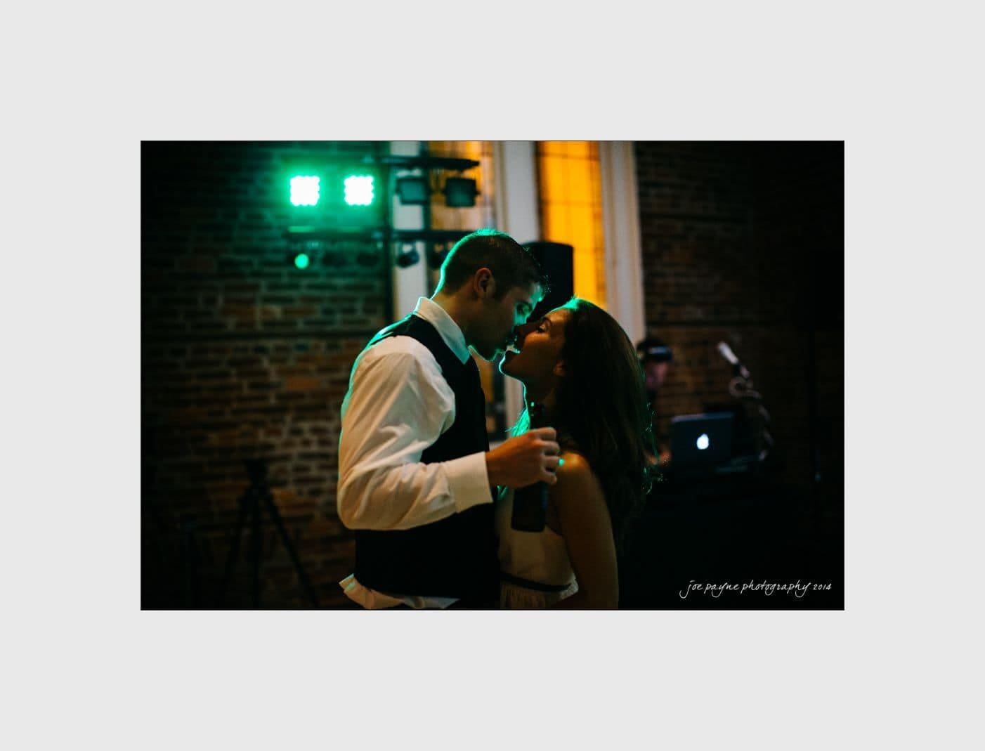 wilmington wedding photographer - nichole & eric A couple dances closely in a dimly lit room. They are dressed in formal attire, with the man holding a drink. Green lights and a brick wall are visible in the background.