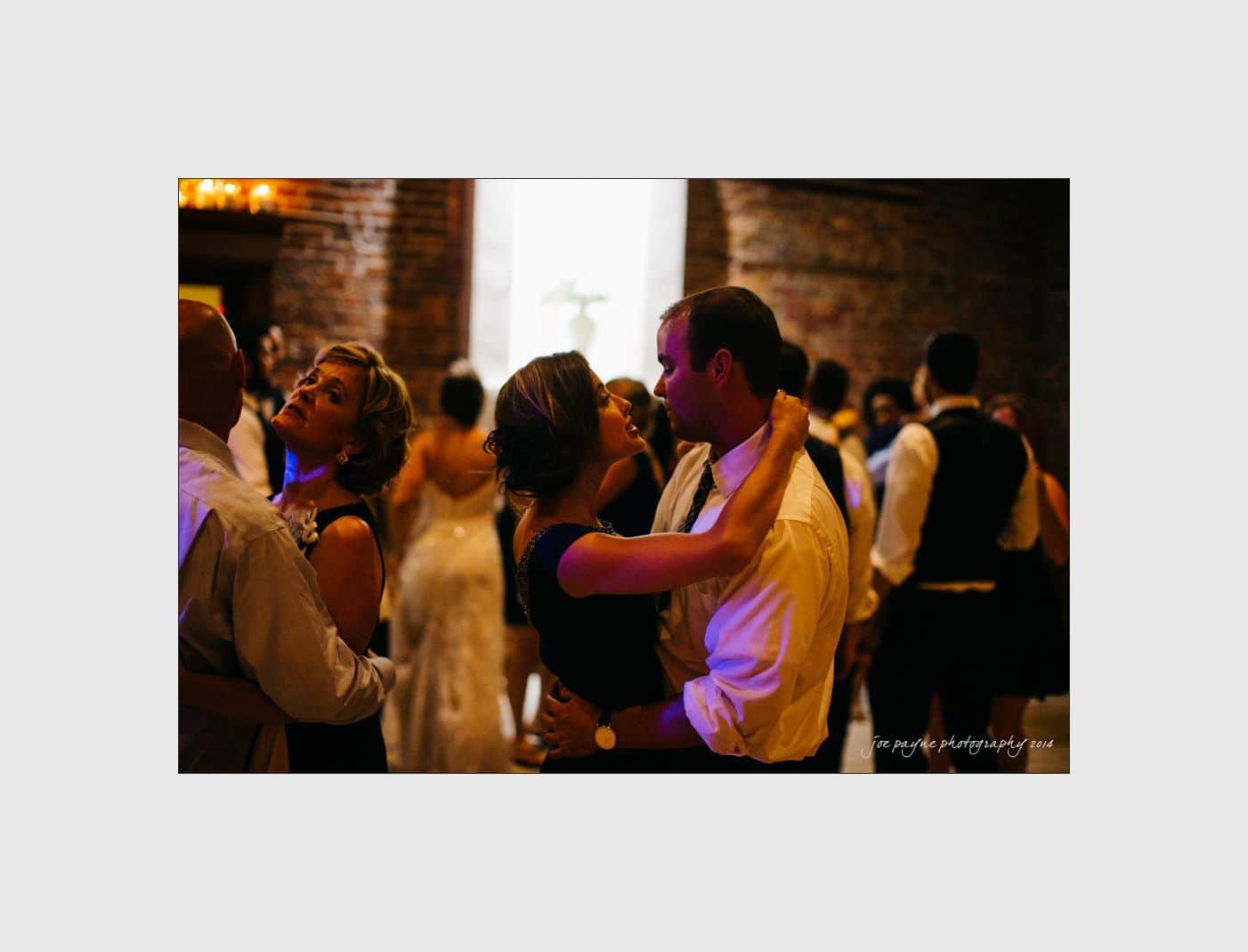 wilmington wedding photographer - nichole & eric Couples dance closely in a dimly lit room with brick walls, dressed in formal attire. A woman wearing a black dress embraces a man in a white shirt as they dance, surrounded by other dancing couples. Warm lighting adds a cozy ambiance.