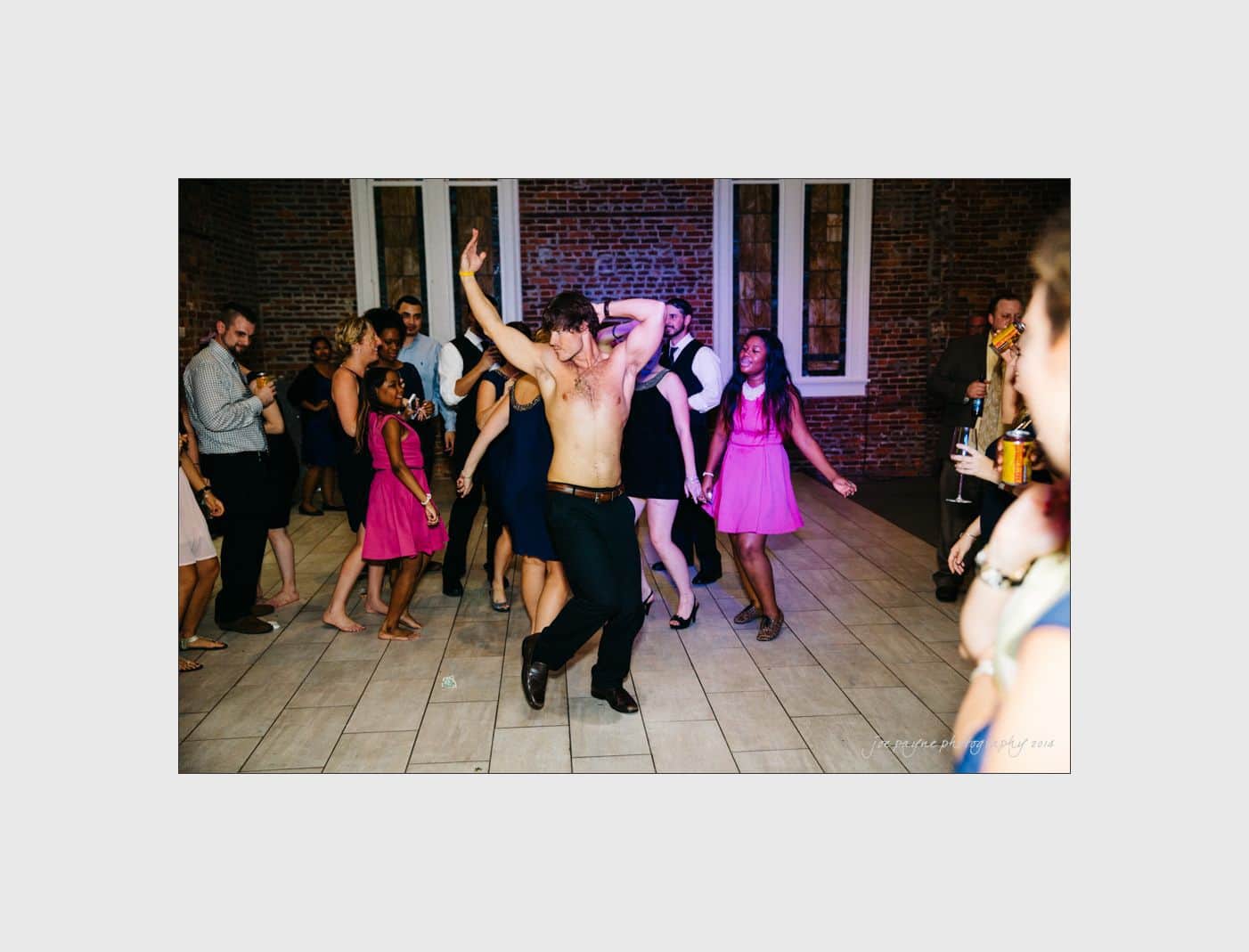 wilmington wedding photographer - nichole & eric Shirtless man dances energetically in a lively, crowded room. People around are dressed in formal attire, some in pink dresses, laughing and enjoying the moment. Brick walls and tall windows are in the background.