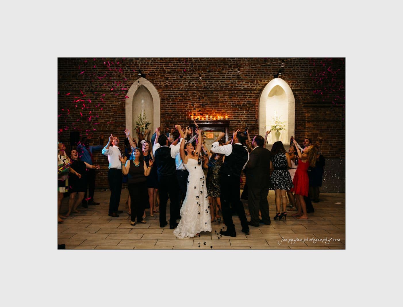 wilmington wedding photographer - nichole & eric A joyful wedding celebration with a bride and groom dancing among guests in an elegant brick-walled venue. Pink confetti fills the air as people clap and cheer around them. The mood is festive and full of happiness.