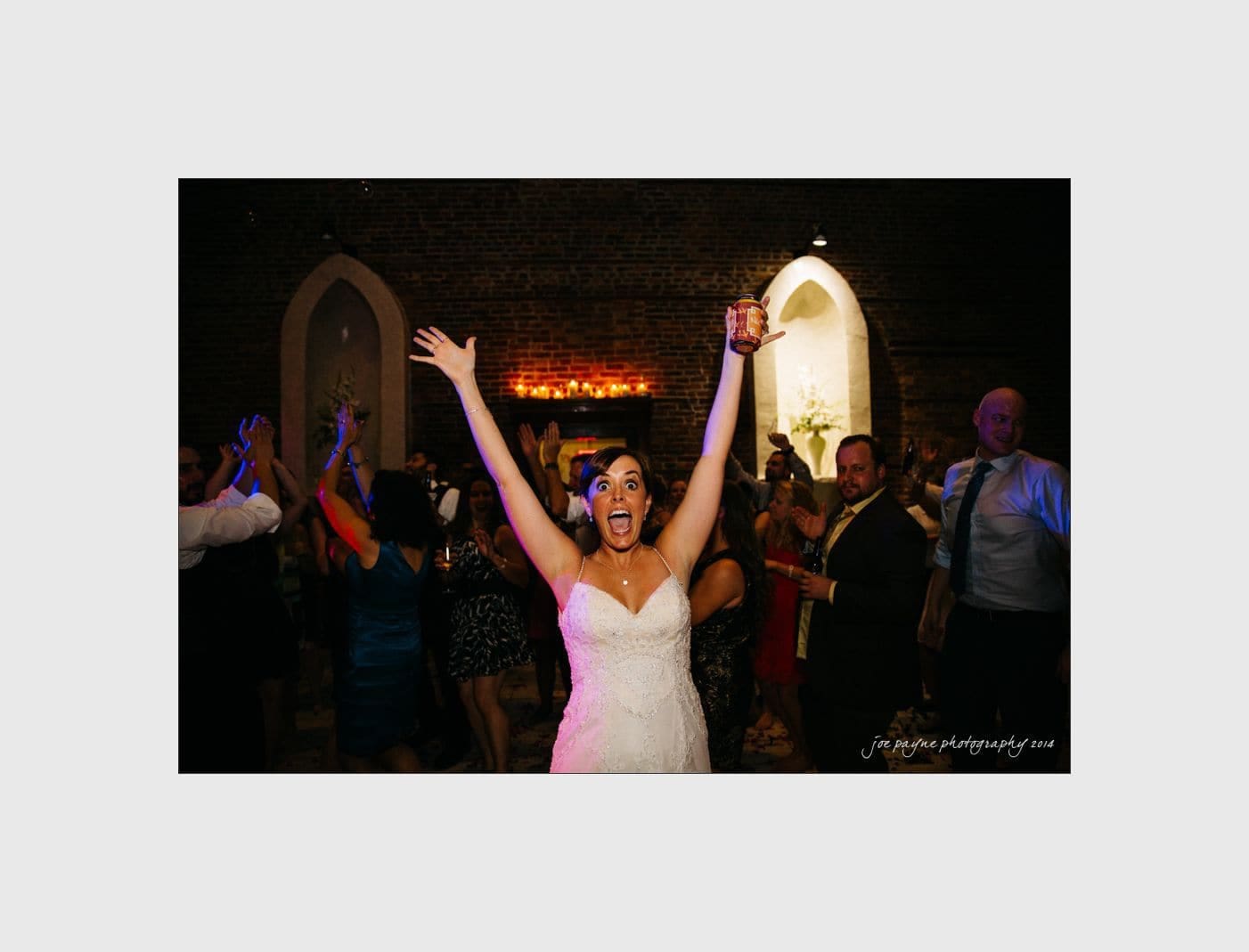 wilmington wedding photographer - nichole & eric A bride in a white dress joyfully raises her arms while holding a drink on a dance floor. Guests dance energetically in the background, and the setting appears to be a dimly lit venue with brick walls.
