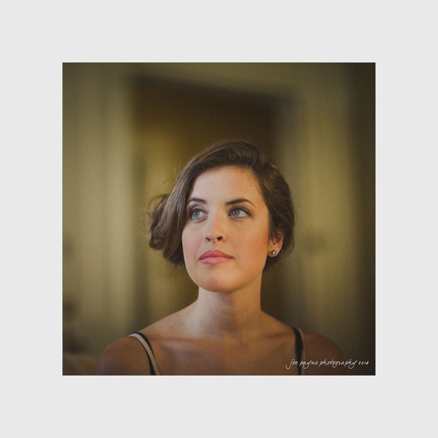 wilmington wedding photographer - nichole & eric A woman with short brown hair and a thoughtful expression looks upward, softly illuminated against a blurred, warm-toned background. Shes wearing a sleeveless top, and the image has a serene, contemplative mood.