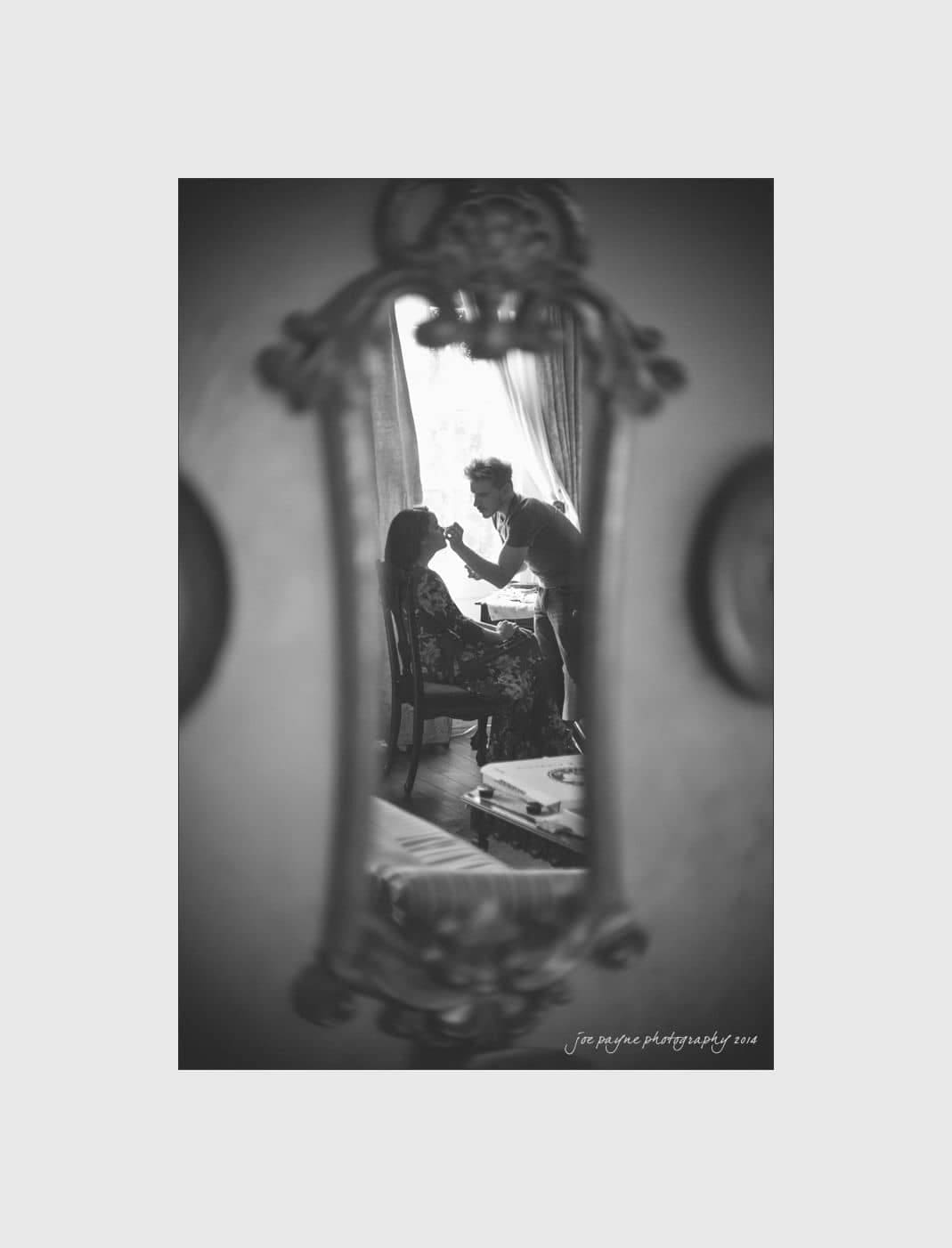 wilmington wedding photographer - nichole & eric A black and white reflection of a makeup artist applying makeup to a seated person, captured in an ornate mirror. The setting appears to be a cozy room with light streaming through a window.