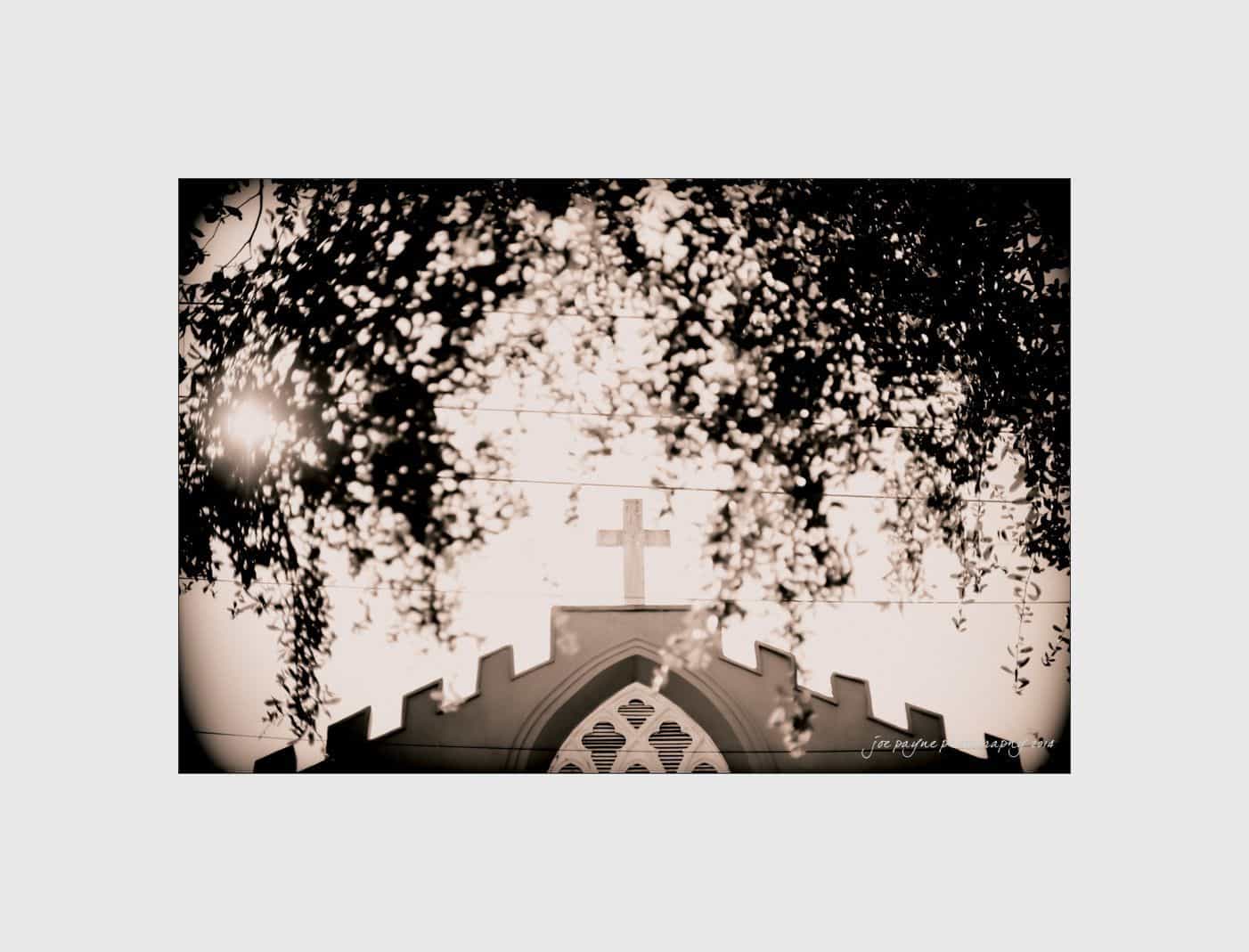 wilmington wedding photographer - nichole & eric A sepia-toned image of a church with a cross on its facade. The sun shines through the branches of a tree, casting shadows on the churchs facade, creating a serene and nostalgic atmosphere.