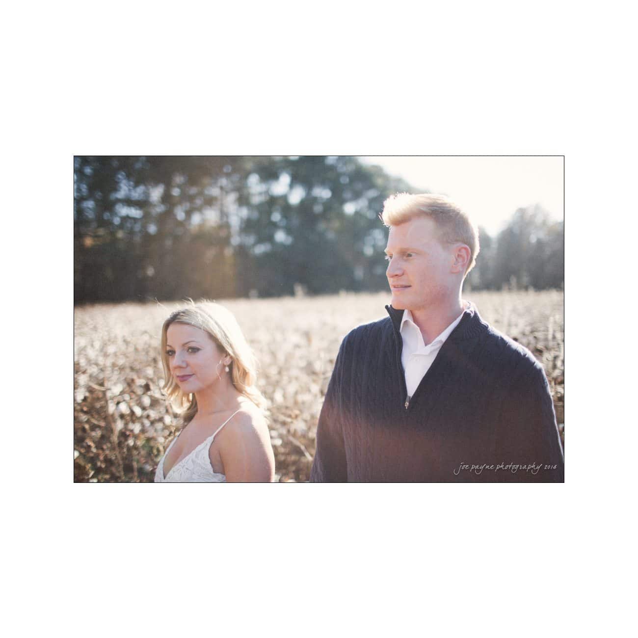 A man and woman stand in a sunny field, surrounded by tall grasses. The woman is in a white dress and the man is in a black sweater. Trees are visible in the background. The lighting is warm and natural.
