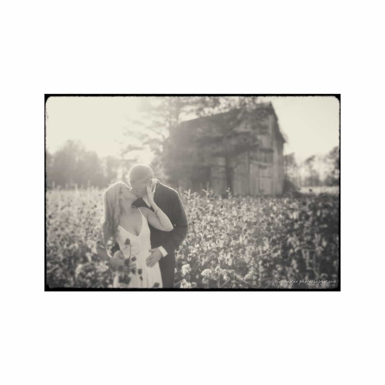 A couple embraces in a field of flowers with an old barn in the background. The image is black and white with a vintage, dreamy effect, showcasing a moment of intimacy and romance.